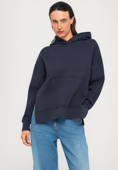Young person wearing a navy oversized hoodie with front pocket and light blue wide-leg jeans, standing against a plain light background.