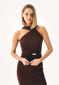 Ruda, be rankovių suknelė su mazgo detalėmis ties iškirpte; modelis laiko juodą tekstūruotą delninę su auksine sagtimi.