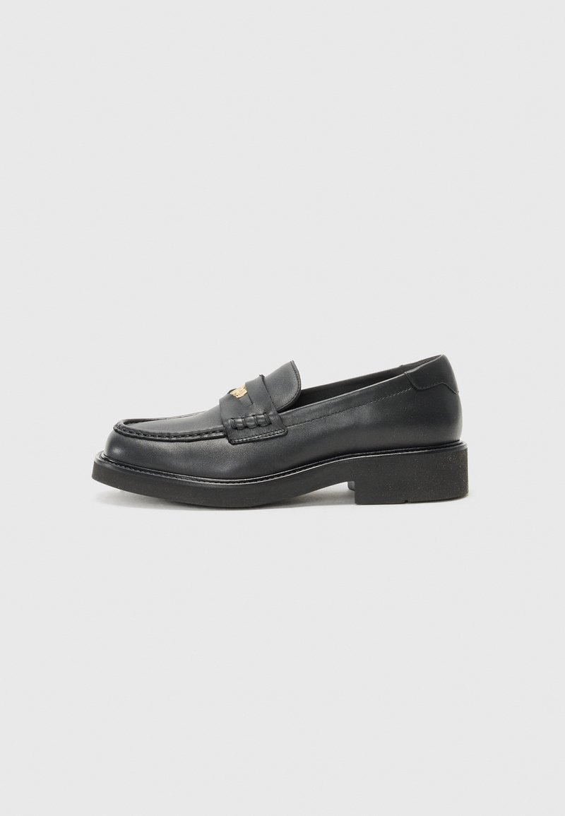 Mocassin en cuir noir avec petit talon carré et emblème doré sur la bride, présenté de profil sur un fond clair uni.