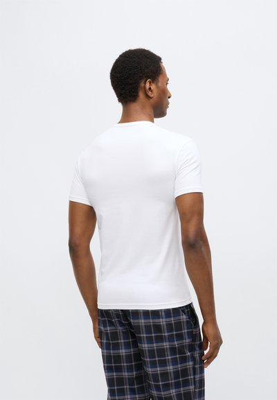 Bílé tričko s krátkými rukávy a kulatým výstřihem, nošené s kostkovanými pyžamovými kalhotami v modré, šedé a černé barvě. Hladká textura látky.