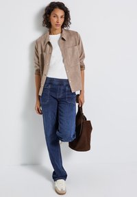 Beige Wildleder-Hemd mit Knöpfen, weißes T-Shirt, dunkelblaue Jeans mit elastischem Bund und cremefarbene Sneakers. Braune Tasche in der Hand, stehend vor einem neutralen Hintergrund.
