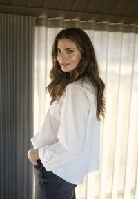 Weiße Bluse mit langen Ärmeln und elastischen Bündchen, kombiniert mit dunklen, hoch taillierten Jeans aus Denim. Der Hintergrund zeigt vertikale, durchsichtige Vorhänge.