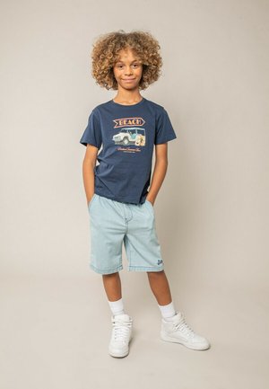 Garçon aux cheveux bouclés debout, souriant, les mains dans les poches de son short bleu clair, portant un t-shirt marine à thème plage et des baskets montantes blanches.