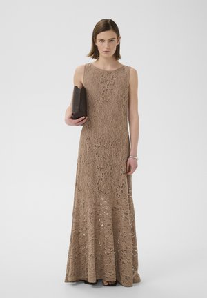 Femme portant une robe longue beige en dentelle sans manches, tenant une pochette noire sur un fond clair uni.