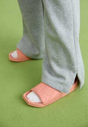 Chinelos de borracha rosa com tiras largas, detalhes texturizados, usados com calças de moletom cinzentas e meias brancas sobre uma superfície verde.
