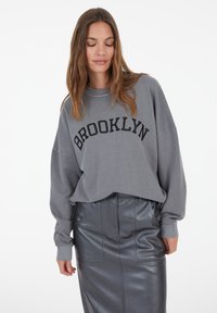 Grey oversized sweatshirt with "BROOKLYN" in black lettering. Paired with a high-waisted grey leather skirt featuring pockets.