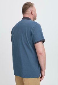 Kék pólóing, textúrált szövettel, rövid ujjakkal és kontrasztos sötét gallérral. Oldalsó hasítékokkal és finom varrás részletekkel az alsó szélén.