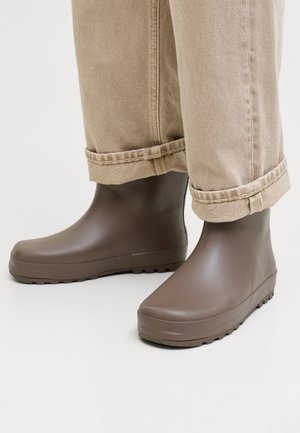 Bottes de pluie en caoutchouc marron pour les chevilles portées avec un pantalon beige retroussé sur un fond clair.