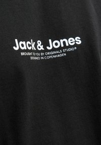 Crna majica s bijelim natpisom. Natpis glasi "Jack & Jones donijet ćete vam Originals Studio® dizajnirano u Kopenhagenu." Glatka tekstura tkanine.