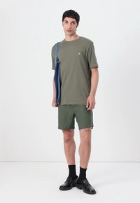 Olivgrünes Baumwoll-T-Shirt, passende Shorts und schwarze Lederschuhe. Blaue Jeans-Tasche mit Schulterriemen. Schlichtes Design, minimale Markenpräsenz.