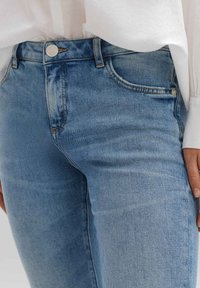 Gros plan d'une personne portant un jean bleu clair et un haut blanc à manches longues, montrant la zone de la taille et une main reposant sur le côté.