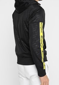 Svart hoodie med ett texturerat mönster, med en gul logosträng längs vänster ärm och en framficka. Justerbar huva ingår.