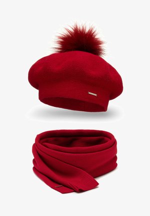 Béret en tricot rouge avec un pompon duveteux sur le dessus, accompagné d'une écharpe rouge assortie. Les deux pièces présentent une texture lisse et une petite étiquette de marque.