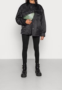 Black satin jacket, green crossbody bag, black leggings, and chunky black boots with buckles. Smooth textures and modern design elements.