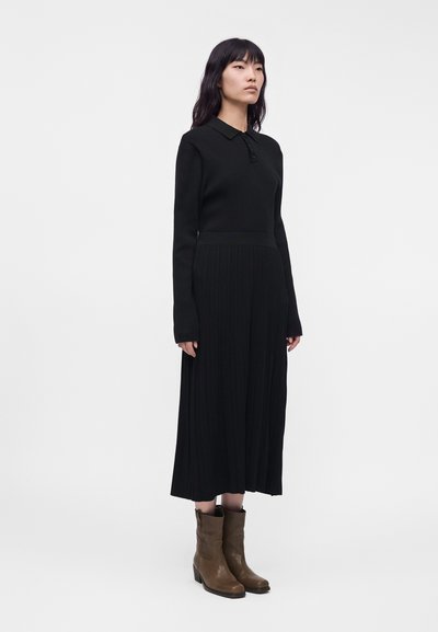 Femme aux longs cheveux noirs portant une robe noire à manches longues et des bottes marron mi-mollet, debout devant un fond blanc uni.