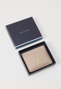 Beige leather cardholder featuring a gold "TH" logo and multiple slots, presented in a navy box with a colorful accent stripe.