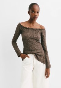 Top marrone off-shoulder con tessuto testurizzato e stropicciato e scollatura arricciata. Maniche lunghe e vestibilità rilassata, abbinato a pantaloni bianchi a gamba larga.