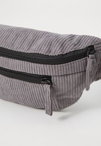 Harmaa corduroy-vyölaukku, jossa on kaksi vetoketjullista taskua, teksturoitu pinta ja mustat vetoketju yksityiskohdat. Kompakti, pyöreä muoto.