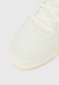 Nahaufnahme der Spitze eines weißen Sneakers mit perforierten Details, strukturierter Naht und weißen Schnürsenkeln vor hellem Hintergrund.