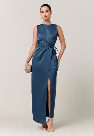 Moteris, vilkinti mėlenu be rankovių satino suknelę su šonine skeltuke, laikanti smėlio spalvos delninę, ją papildo auksiniai auskarai, apyrankė ir subtilios aukštakulnės basutės.