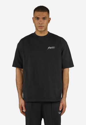 Mladý muž nosiaci čierne nadrozmerné tričko a zodpovedajúce čierne nohavice s malými bielymi nápismi "Pegador" na hrudníku a stehne.