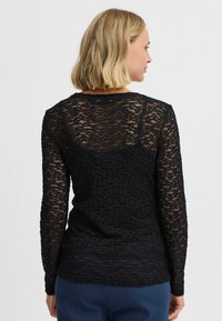 Zwarte top met lange mouwen van kant met een textuurpatroon. Heeft een ronde hals en een aansluitende pasvorm, gedragen over blauwe broek.