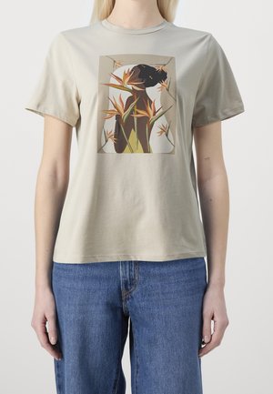 T-shirt beige avec un graphique d'une femme à la peau foncée de profil entourée de fleurs orange d'oiseau de paradis, porté avec un jean bleu.