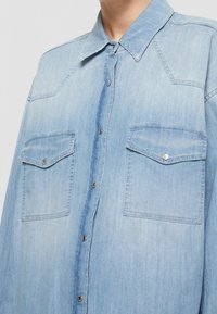 Camisa de mezclilla azul claro con cuello, dos bolsillos en el pecho con cierre de botón y un corte holgado. Textura suave y un sutil desgaste.