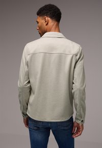 Camisa de manga larga gris claro con cuello, que presenta una textura suave y puños abotonados, usada sobre unos jeans azules.