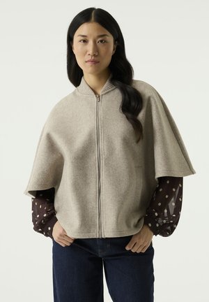 Woman with long dark hair wears a beige zip-up cape over a brown polka dot top and dark jeans, standing with hands in pockets.
