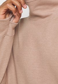 Sudadera de cuello vuelto color marrón claro con mangas largas, fabricada en un tejido suave y liso, con un corte relajado; detalle de costura mínima.