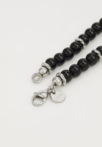 Chaîne de perles noires avec accents en métal argenté, dotée d'un fermoir en forme de homard et d'une étiquette circulaire. Texture lisse avec des finitions alternant poli et mat.