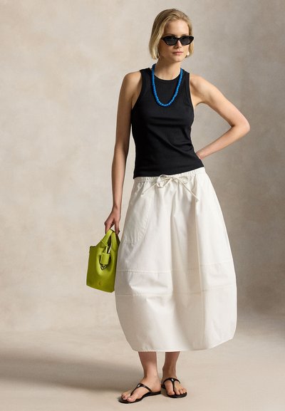 Γυναίκα με μαύρο αμάνικο τοπ, λευκή φούστα, μαύρα σανδάλια, μπλε κολιέ, γυαλιά ηλίου, κρατώντας μικρή πράσινη τσάντα, στέκεται μπροστά σε ουδέτερο φόντο.