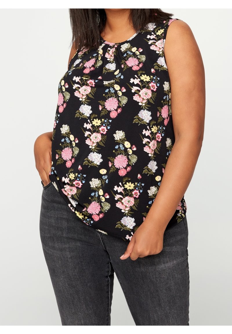 Femme portant un haut noir sans manches avec un motif floral rose, blanc et jaune, associé à un jean gris foncé, mains dans les poches.