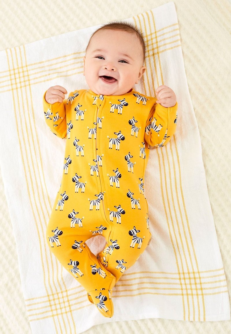 Gul baby-suit med sort og hvid zebra mønster, lange ærmer og lynlås foran, lagt på et hvidt tæppe med gule striber.