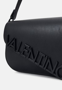 Svart läder axelremsväska med en magnetisk klaff, prydd med upphöjt "Valentino"-logotyp i en glänsande finish, samt en texturerad yta.