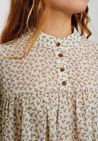 Blouse florale en tissu crème, avec un petit col, des boutons en bois, et une jupe froncée ornée de fleurs violettes et de feuilles vertes.