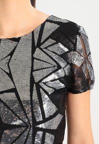 Svart topp med ett geometriskt mönster gjort av silverpaljetter. Korta, genomskinliga ärmar och textil med struktur förstärker det visuella intrycket.