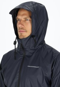 Donkerblauwe jas met capuchon, voorzien van een ritssluiting en verstelbare trekkoorden. Materiaal lijkt waterdicht met een gladde textuur. Merkennaam zichtbaar.