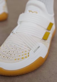 Λευκό αθλητικό παπούτσι με κίτρινες λεπτομέρειες, υφασμάτινη κορυφή με υφή, δύο φαρδιές διασταυρούμενες ζώνες και σόλα από καουτσούκ γιούμ σε γκρι επιφάνεια.