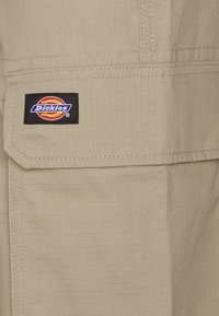 Tan werkbroeken van duurzaam stof hebben een grote voorzak met een zwart Dickies-logo patch en zichtbare stikdetails.