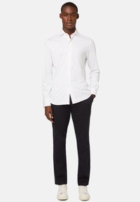 Valkoinen pitkähihaisella nappipaidalla, jossa on kaulus, yhdistettynä tummiin housuihin ja valkoisiin lenkkareihin, klassinen malli ja sujuva rakenne.