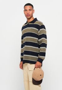 Gestreepte longsleeve polo in olijfgroen, marineblauw en wit. Bevat een kraag en een logo op de borst. Houdt een beige en bruine pet vast.