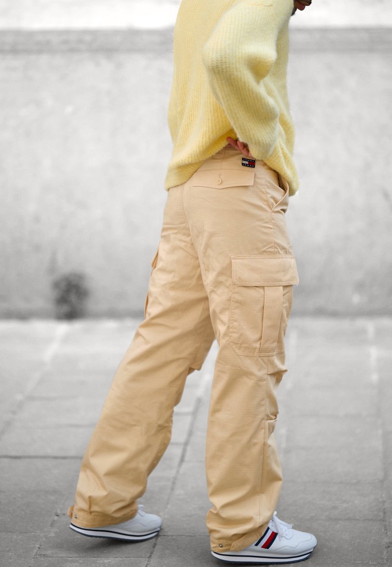 Personne portant un pantalon cargo beige, des baskets blanches avec des bandes rouges et bleues, et un pull jaune, debout sur un trottoir pavé.