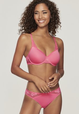 Lächelnde Frau mit lockigem Haar, die einen rosa Spitzen-BH und passende Unterwäsche trägt, vor hellem Hintergrund stehend.