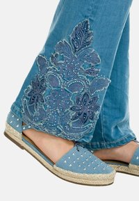 Denim espadrilles dengan kain biru, dihiasi dengan stud perak, dan sol jute bergaya espadrille. Bordir bunga pada celana jeans panjang pergelangan kaki.