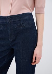 Pantalones de mezclilla oscuros de cintura alta con una textura suave, dos bolsillos delanteros y detalles de costura visibles. Combinados con una blusa de color rosa claro.