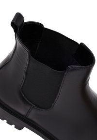 Stivale alla caviglia in pelle nera con pannello elastico laterale, texture liscia, punta rotonda e suola robusta per trazione. Fettuccia per indossarlo facilmente.