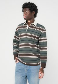 Ung mann med vitiligo iført en grønn, brun og hvit stripete langermet polo-skjorte og lyseblå jeans, som står med den ene hånden i lommen.