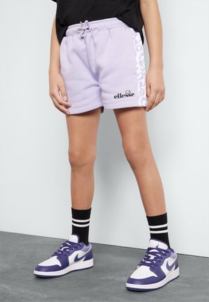 Osoba nosząca jasnofioletowe spodenki ellesse z paskiem w cętki, czarne skarpetki z białymi paskami oraz fioletowo-białe sneakersy Nike.